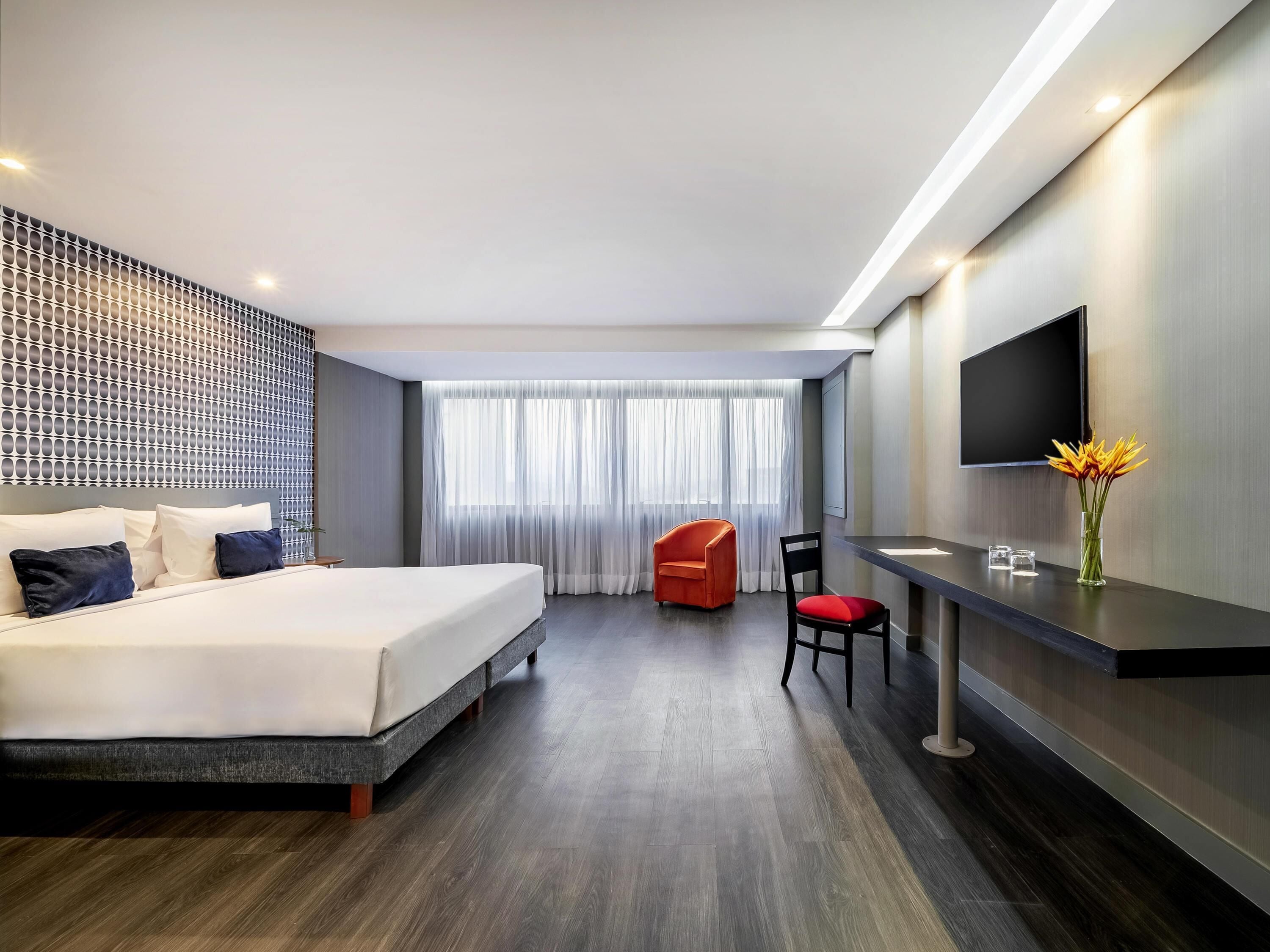 Una habitación de hotel moderna con una cama, un escritorio, una silla, un televisor y un amplio ventanal con cortinas.
