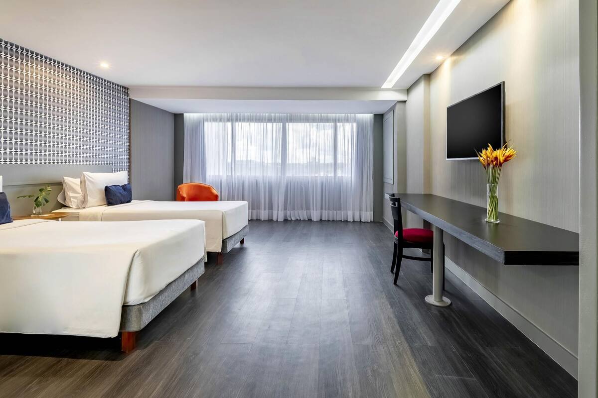 Ein Hotelzimmer mit zwei Betten, einem Schreibtisch, einem Fernseher und einem großen Fenster mit Vorhängen.