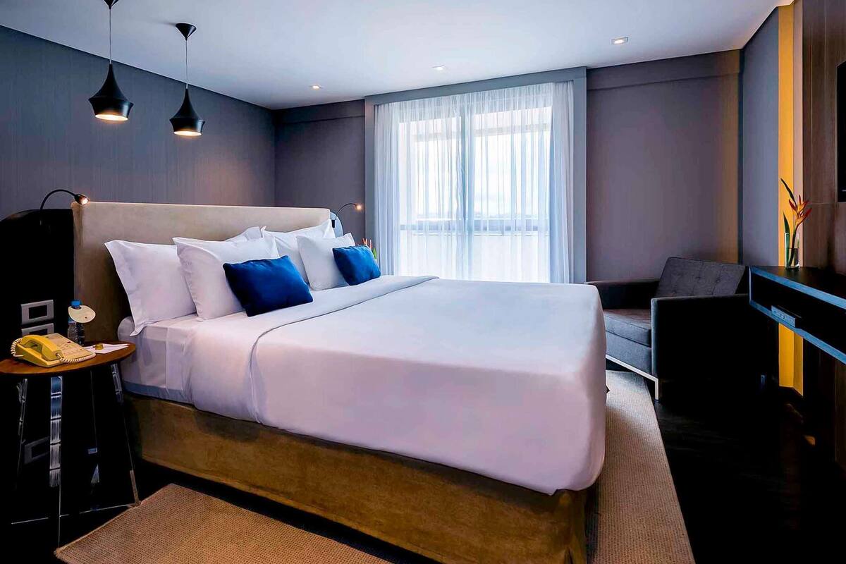 Ein modernes Hotelzimmer mit einem großen Bett, einem Nachttisch, einer Lampe und Blick auf die Stadt durch das Fenster.