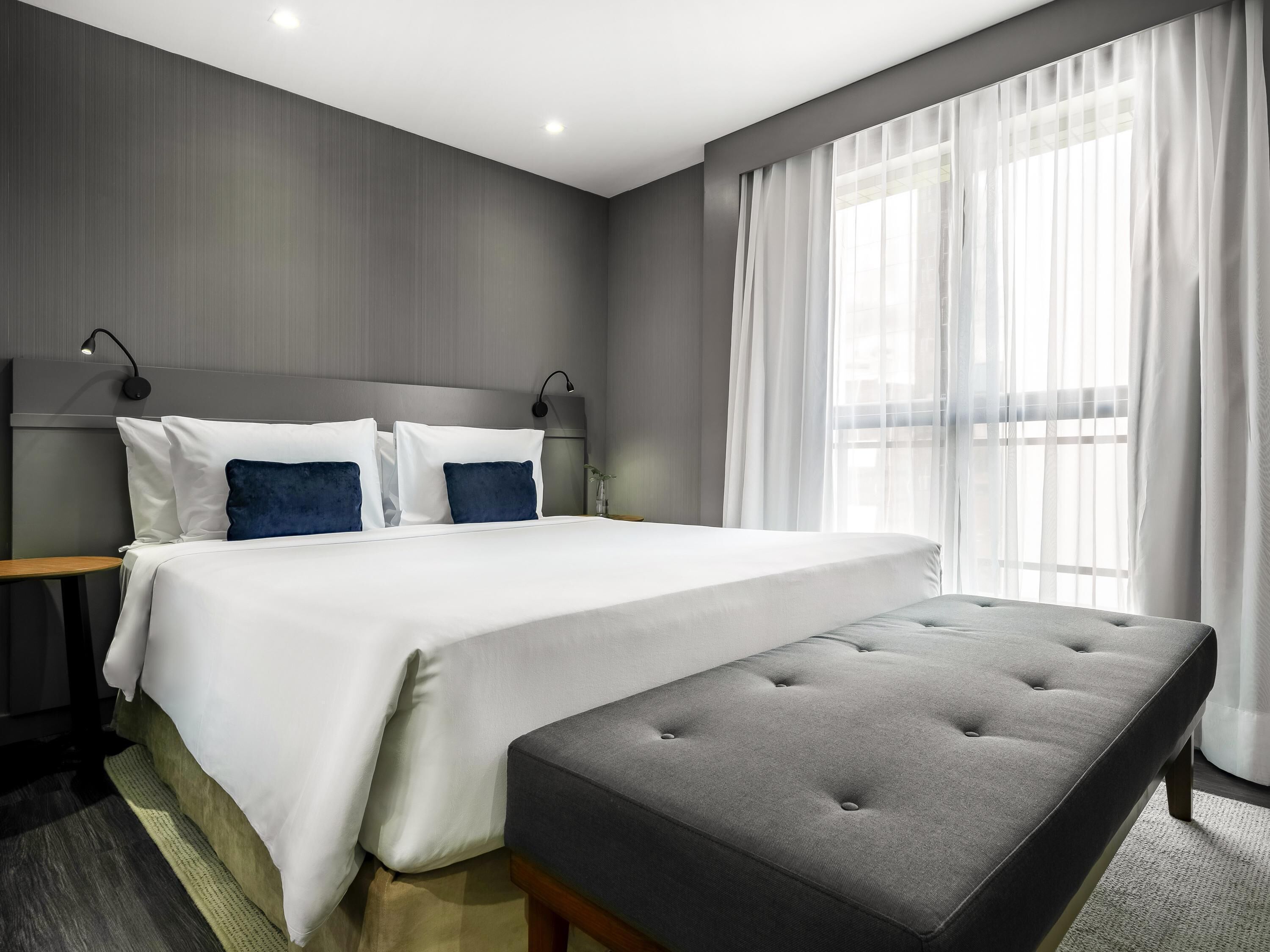 Una habitación de hotel moderna con una cama grande, una mesita de noche, una lámpara y una ventana con cortinas translúcidas.