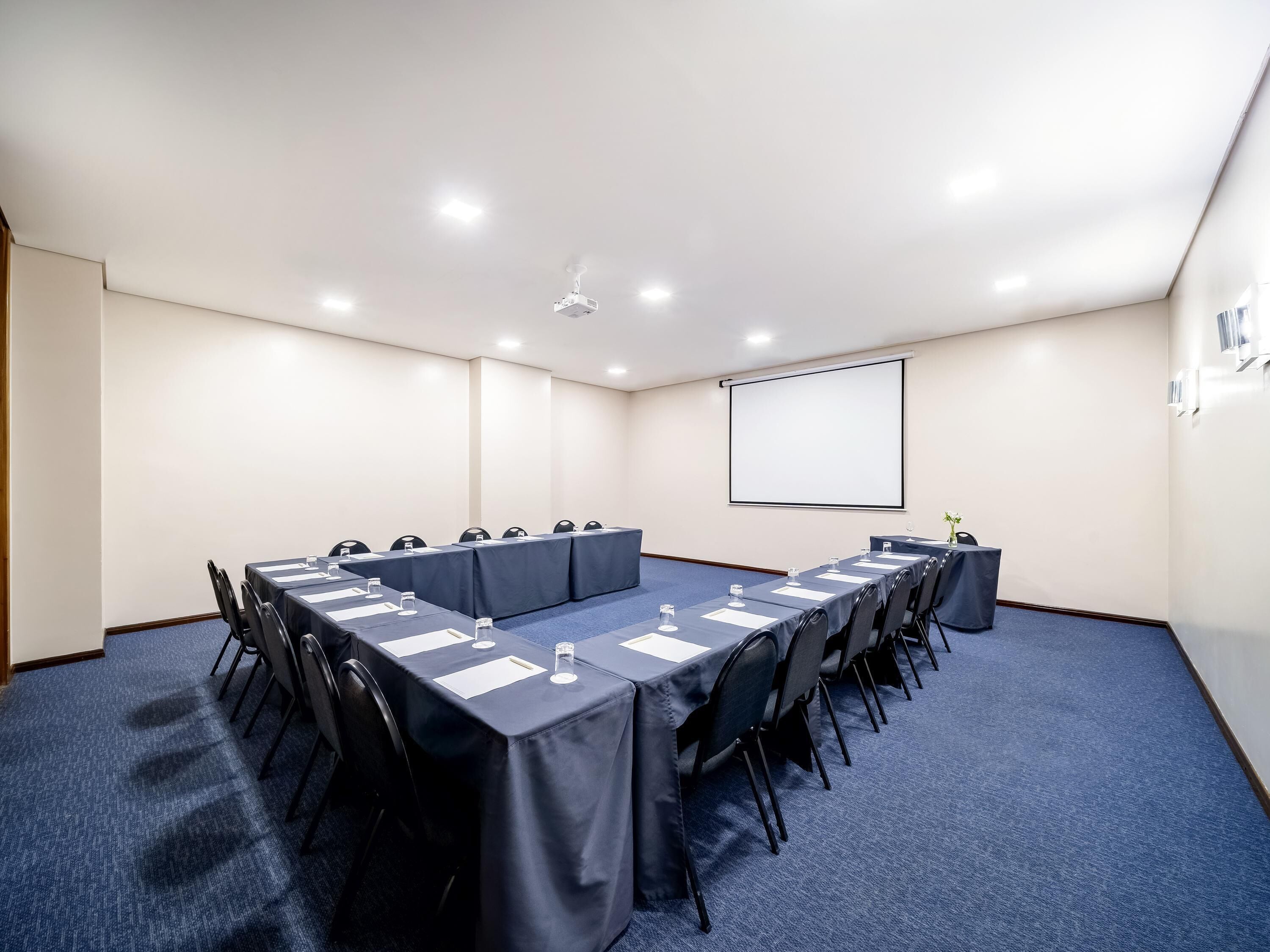 A conference room with a long table, chairs, a projector screen, and a carpeted floor.