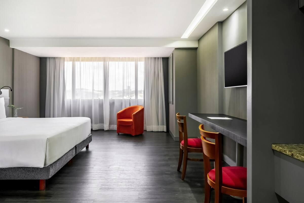 Ein Hotelzimmer mit Bett, Schreibtisch, zwei Stühlen, Fernseher und einem Fenster mit Vorhängen.