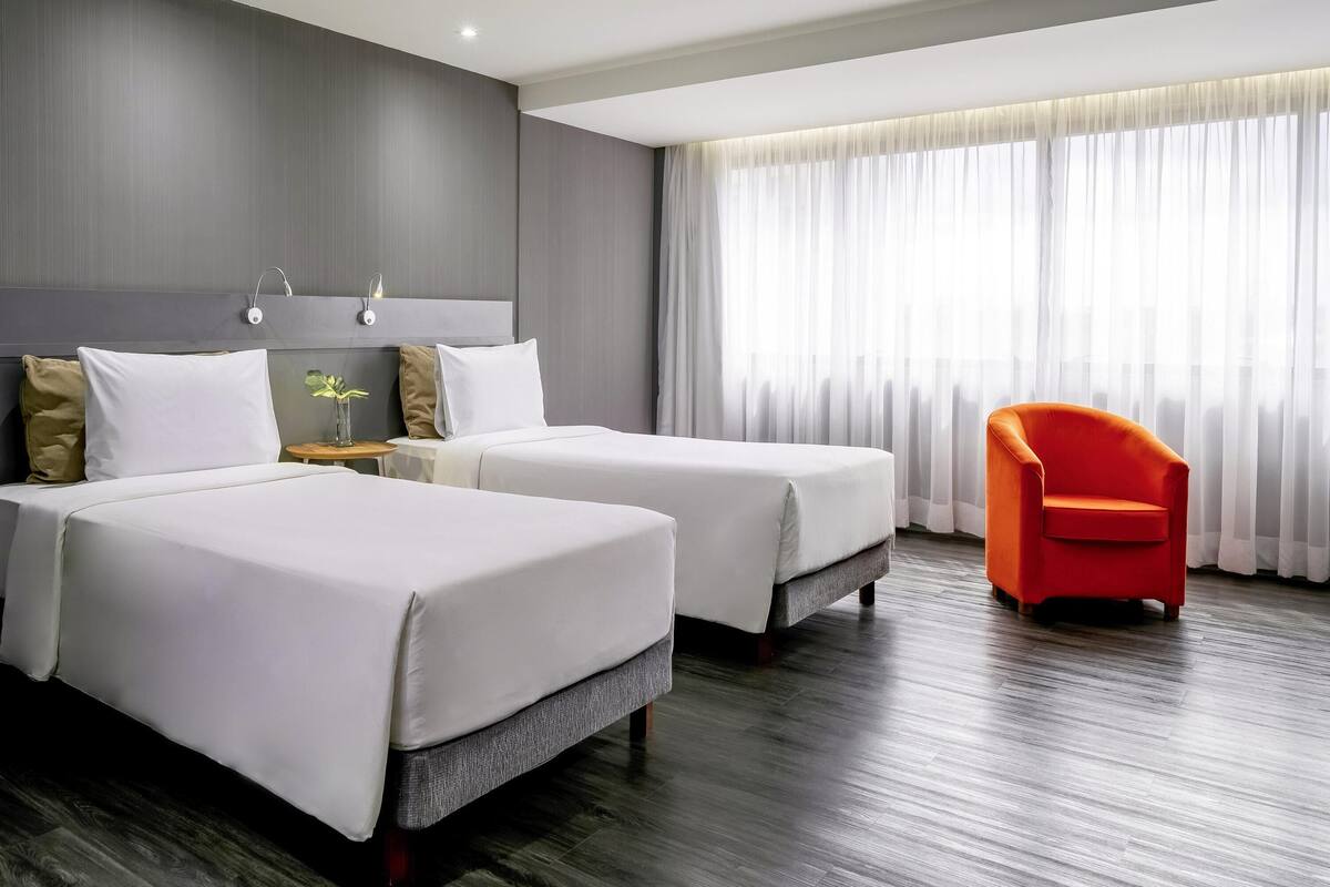 Ein Hotelzimmer mit zwei Betten, einem Nachttisch, einem Sessel und einem großen Fenster mit durchscheinenden Vorhängen.