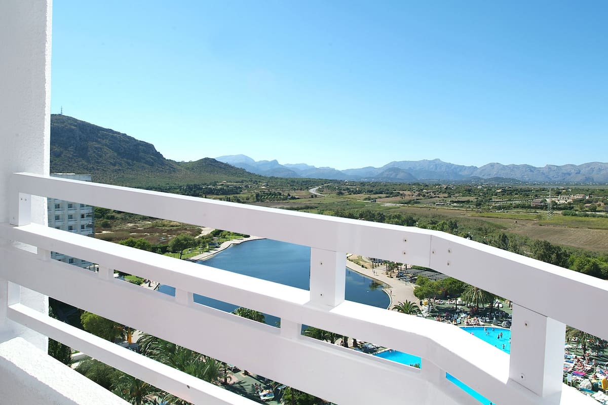 Un balcón con barandal blanco que da a un paisaje pintoresco, con un río, montañas y una zona de piscina.