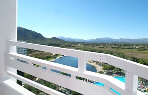 A balcony with a white railing overlooking a scenic landscape, including a river, mountains, and a pool area.