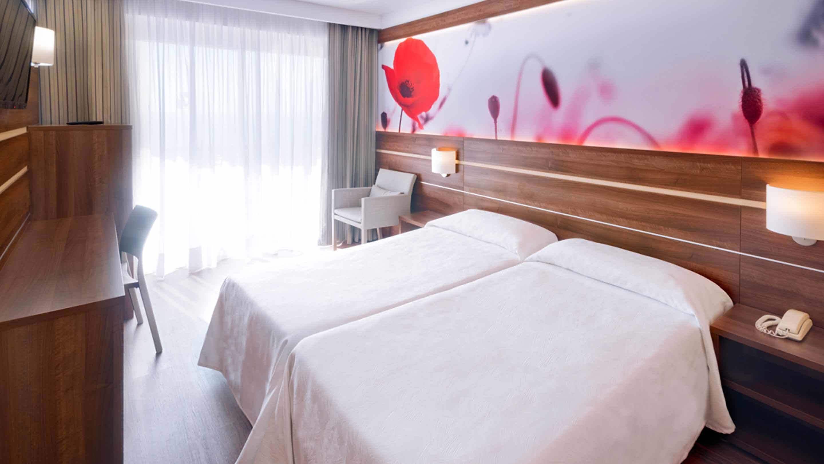 Hotellihuone, jossa on kaksi sänkyä, työpöytä, tuoli, puhelin ja seinä, joka on koristeltu kukkakuvioisella seinämaalauksella.