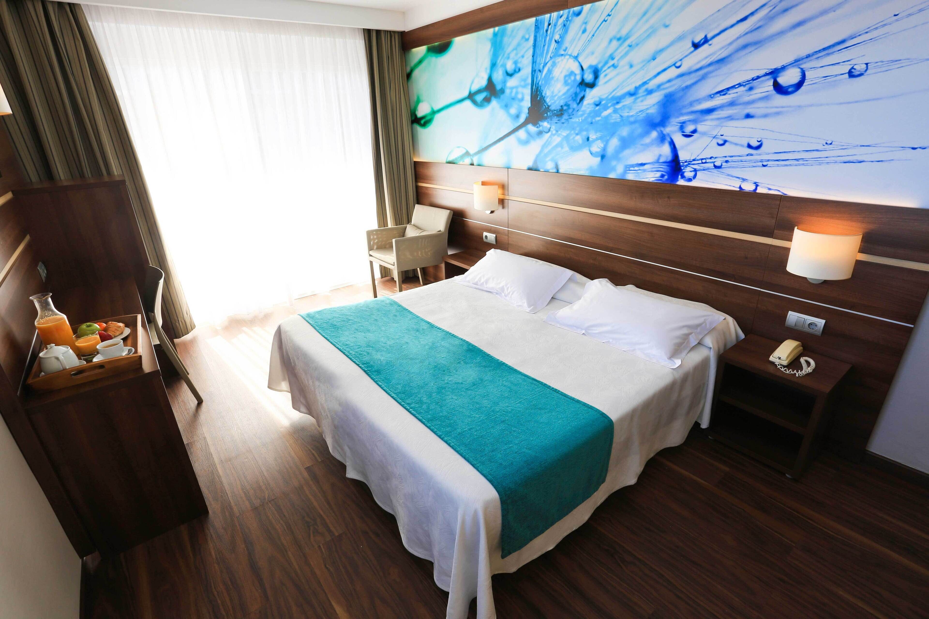 A hotel room with a bed, a bedside table with a lamp, a chair, a wooden wardrobe, and a wall with a blue abstract design.