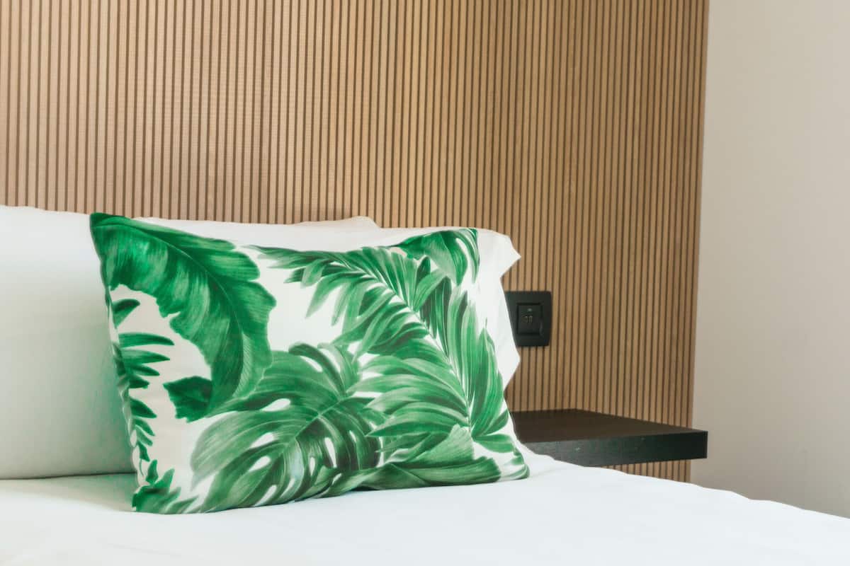 A hotel room with a bed, a pillow with a green leaf pattern, a wooden panel wall, and a black nightstand.