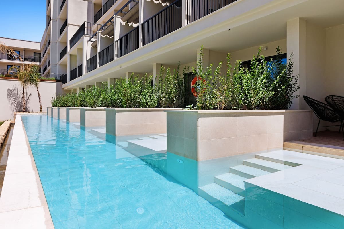 A swimming pool with steps leading into it, surrounded by greenery and a building with balconies.