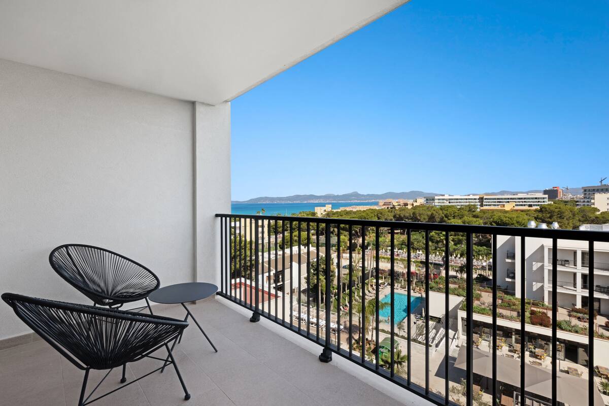 A balcony with two black wireframe chairs and a small table, overlooking a pool and buildings.