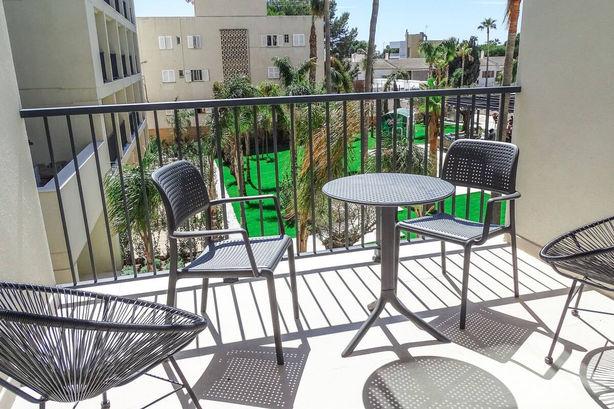 A balcony with two chairs and a table, overlooking a green garden and buildings.