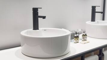 A modern bathroom with a white sink, black faucet, and a mirror reflecting the sink. There are small bottles and a glass on the counter.