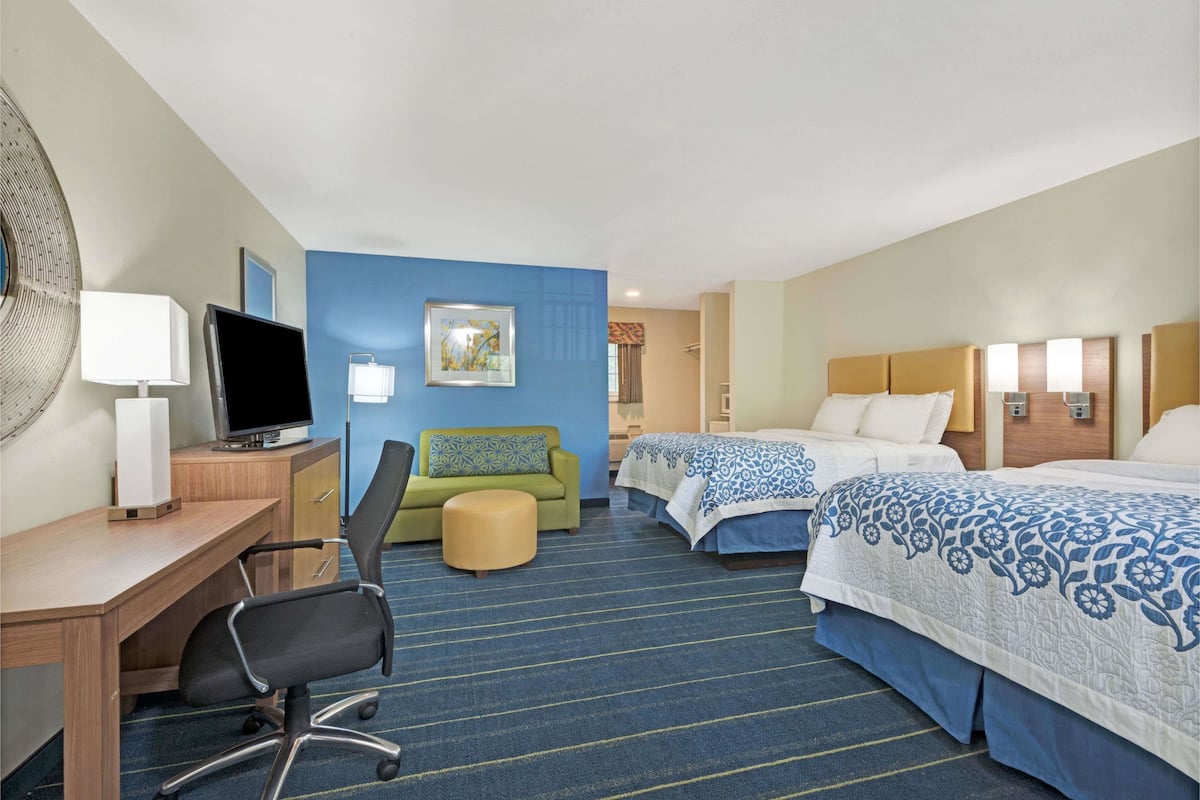 A hotel room with a desk, two beds, a green sofa, and a large window.