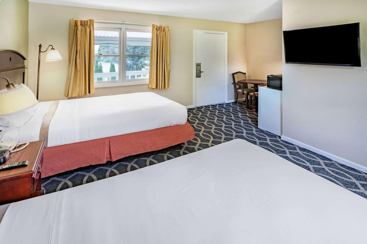 Ein Hotelzimmer mit zwei Betten, einem Schreibtisch mit Lampe, einem Fernseher und einem Fenster mit Vorhängen.