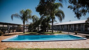 A rectangular swimming pool with a brick patio, surrounded by palm trees and a building with a metal roof. - Kalgoorlie Overland Motel (Somerville)