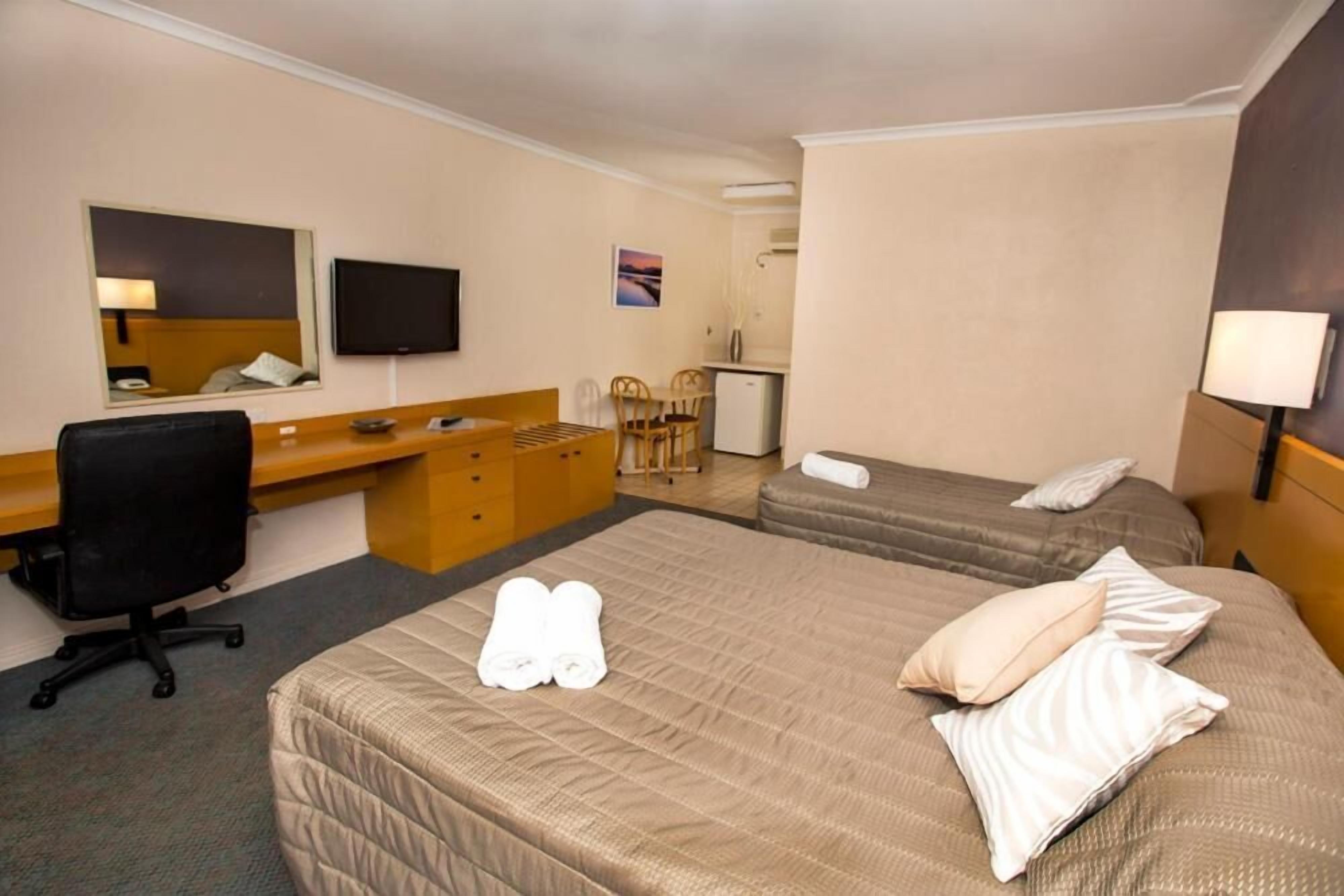 A hotel room with two beds, a desk, a chair, a television, and a small kitchenette.