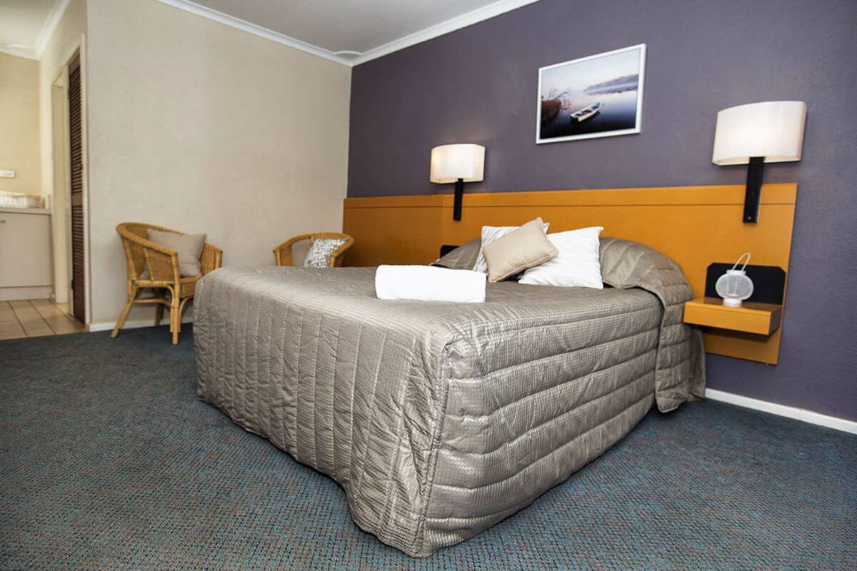 Habitación de hotel con cama, dos lámparas de pared, una mesita con lámpara, una silla y un cuadro en la pared.