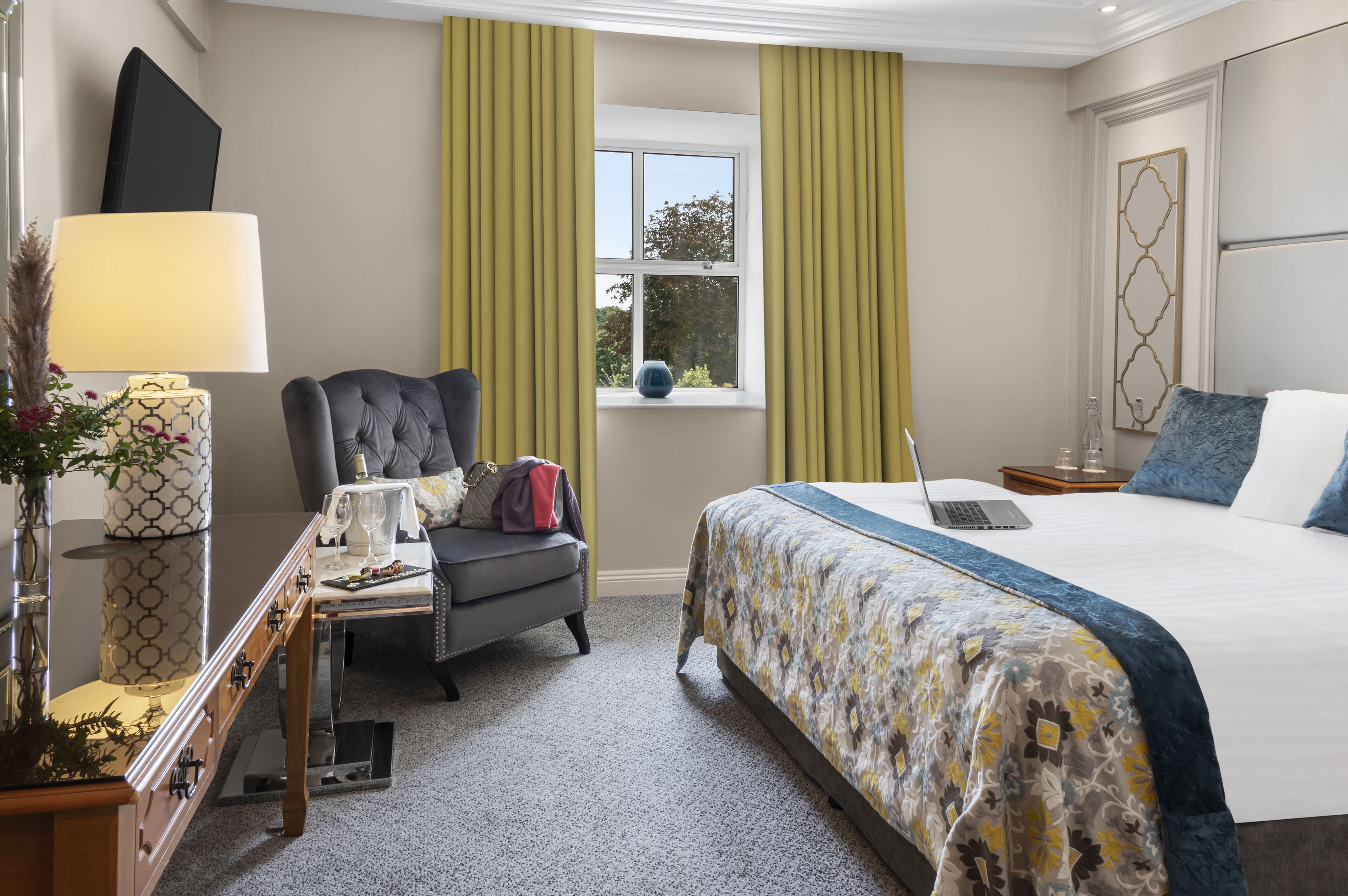 A hotel room with a bed, a desk with a lamp, a chair, a window with curtains, and a view of the outdoors.