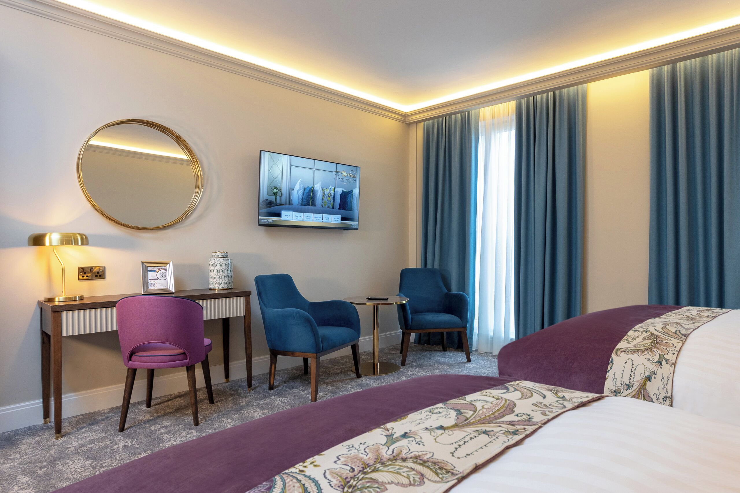 A hotel room with a large bed, a desk with two chairs, a round mirror, a flat-screen TV, and a window with blue curtains.