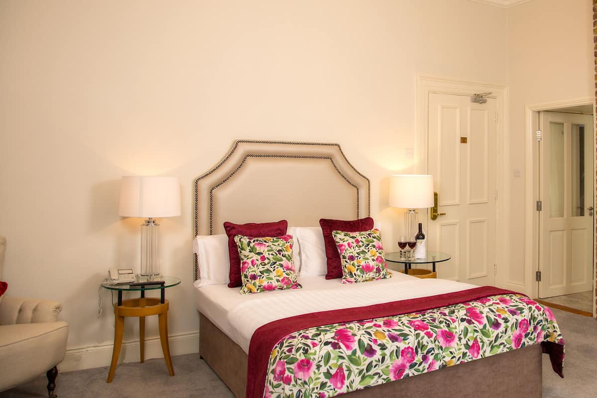 A hotel room with a bed, two bedside lamps, a television mounted on a brick wall, and a fireplace.