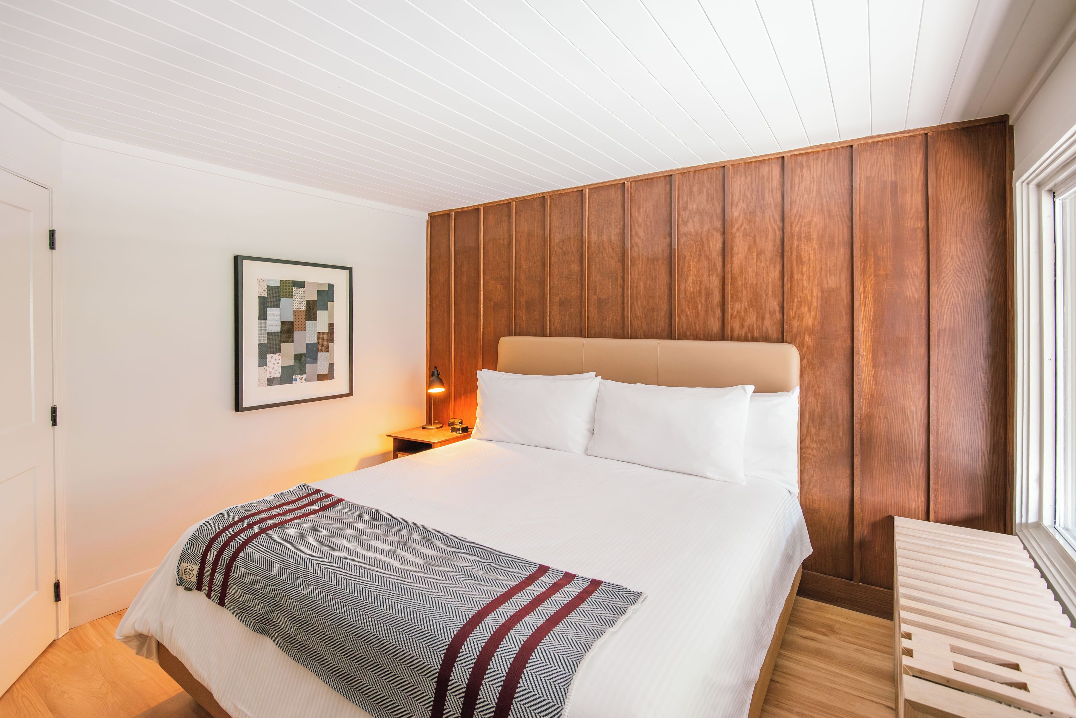 A neatly arranged bedroom with a large bed, a wooden headboard, a bedside table with a lamp, and a framed artwork on the wall.