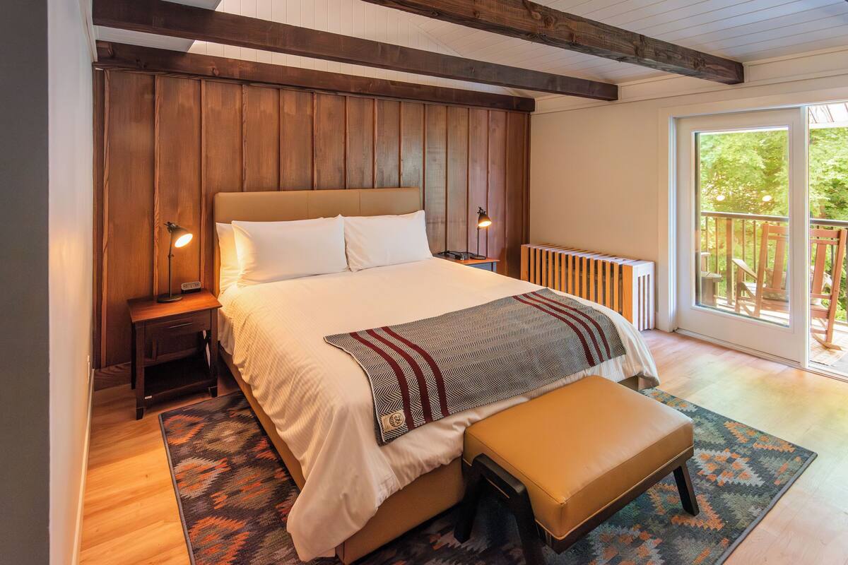 A bedroom with a large bed, a bench, a nightstand, a lamp, and a view of the outdoors.
