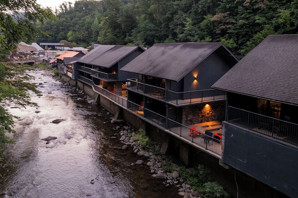 Filas de cabañas modernas con techos oscuros y balcones con vista a un río, rodeadas de un bosque denso.