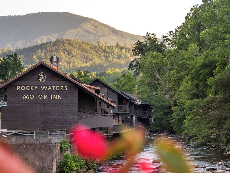 Una locanda con uno sfondo montuoso, con un cartello ben visibile e un fiume in primo piano.
