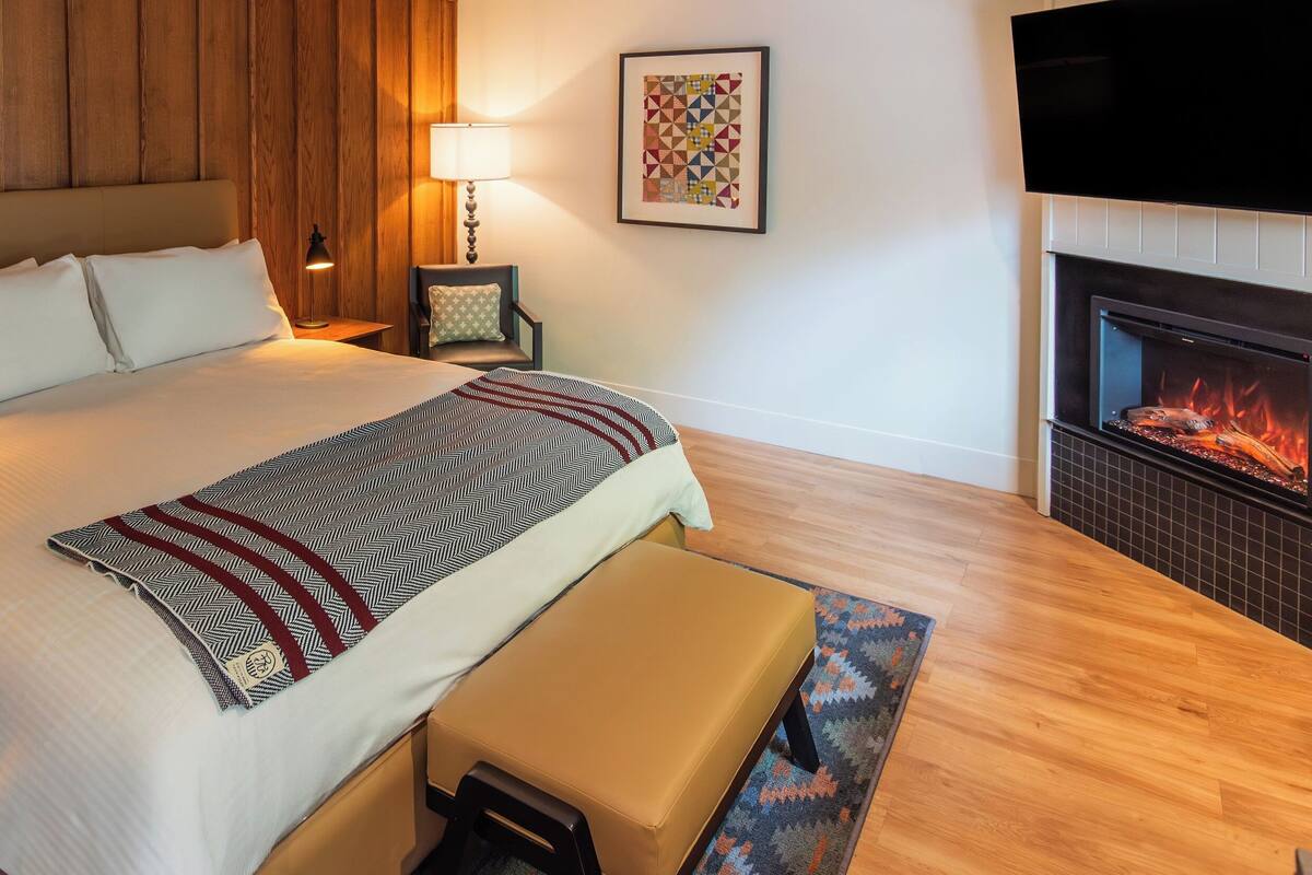 A hotel room with a bed, a bench, a fireplace, a TV, and a lamp.