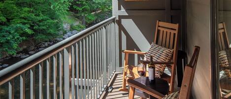 A wooden deck with a railing, two wooden chairs with patterned cushions, and a table with a bottle and glass.