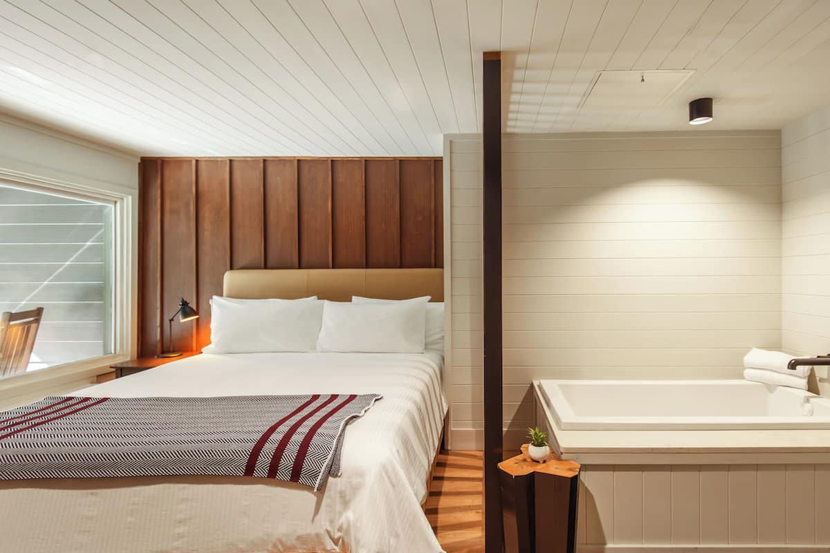 A modern hotel room with a large bed, a wooden headboard, a built-in bathtub, and a window with blinds.