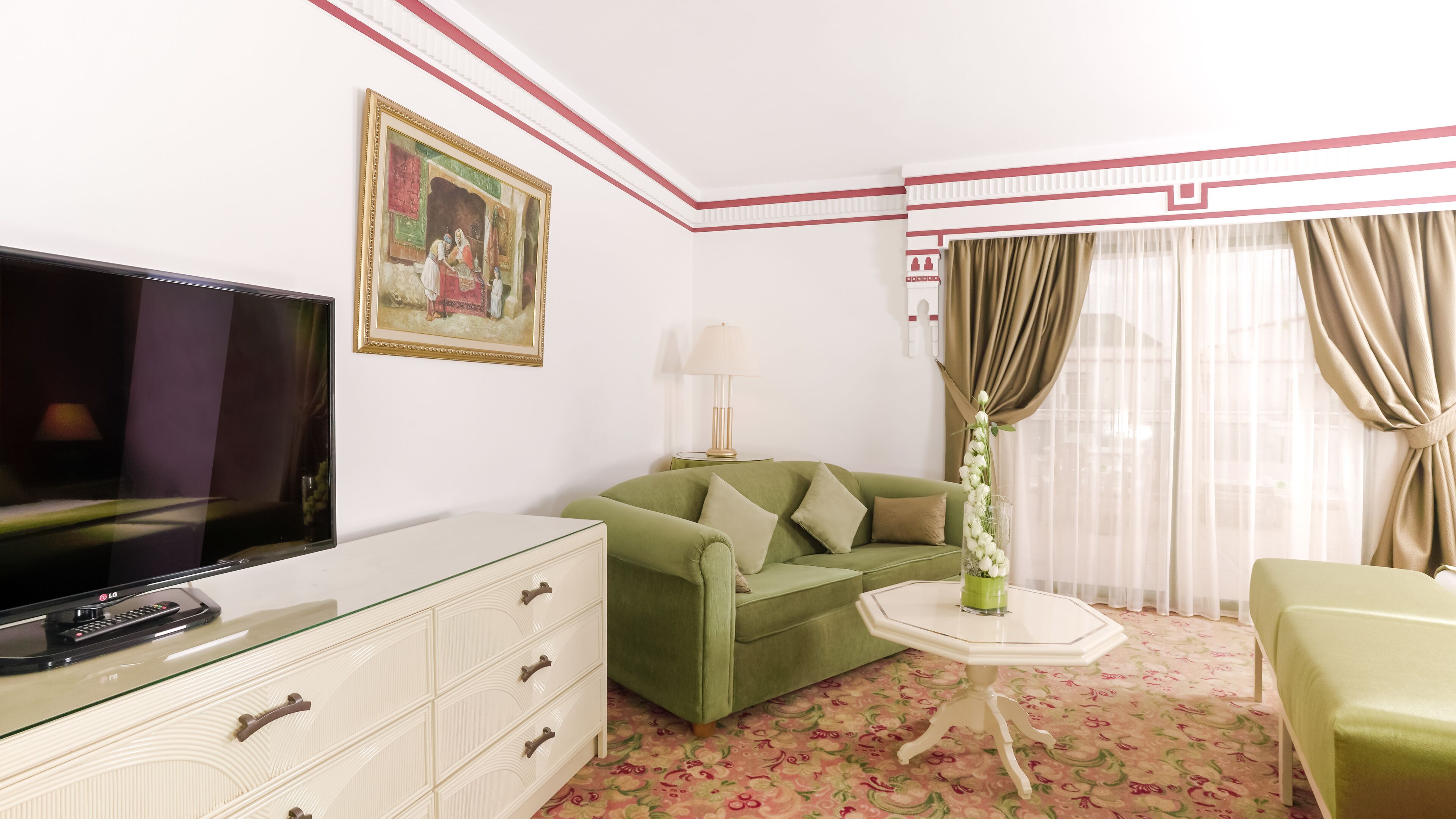Habitación de hotel con sofá verde, televisor de pantalla plana, una mesita con lámpara y alfombra estampada.