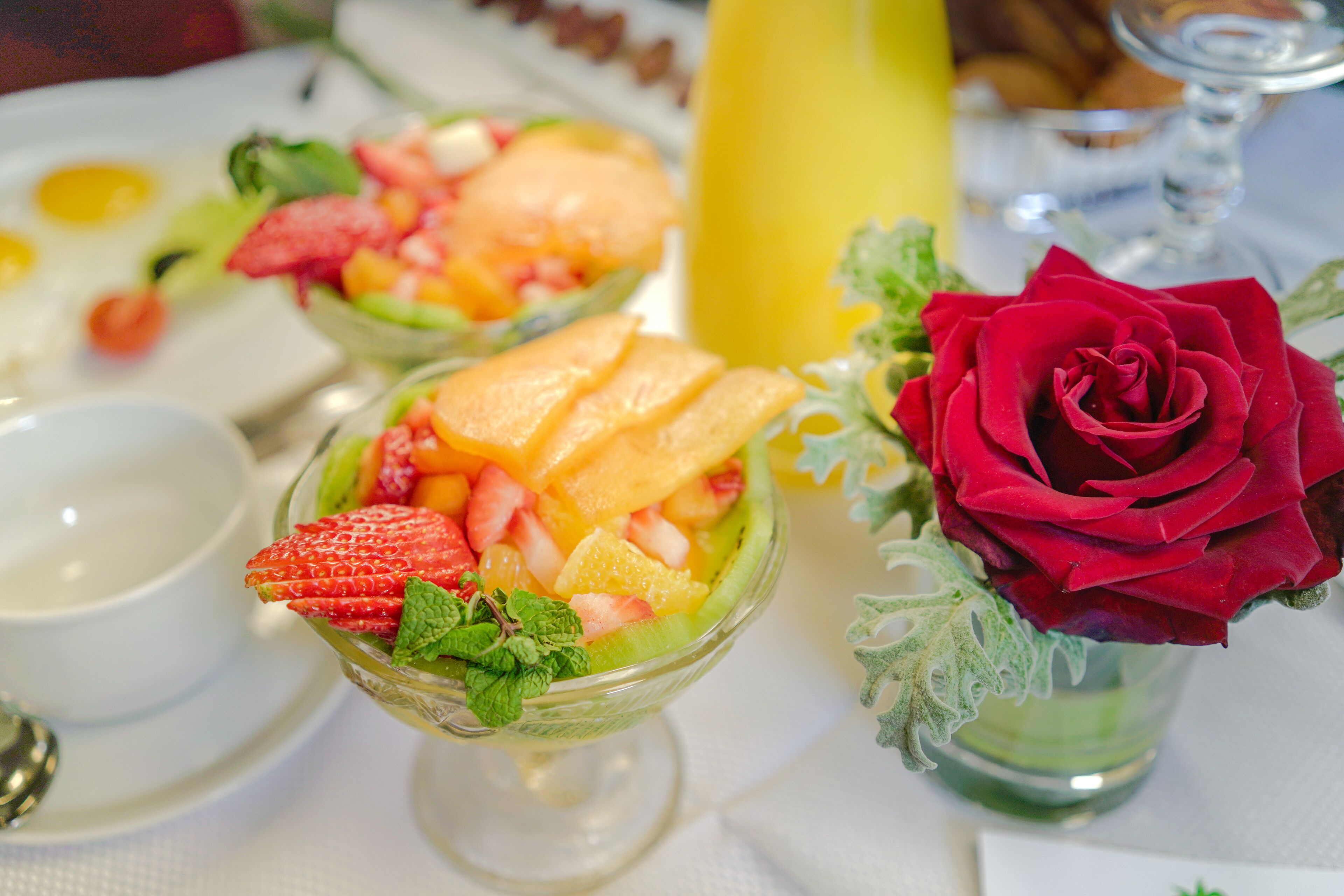Una mesa puesta con una ensalada de frutas, una rosa y un vaso de jugo.