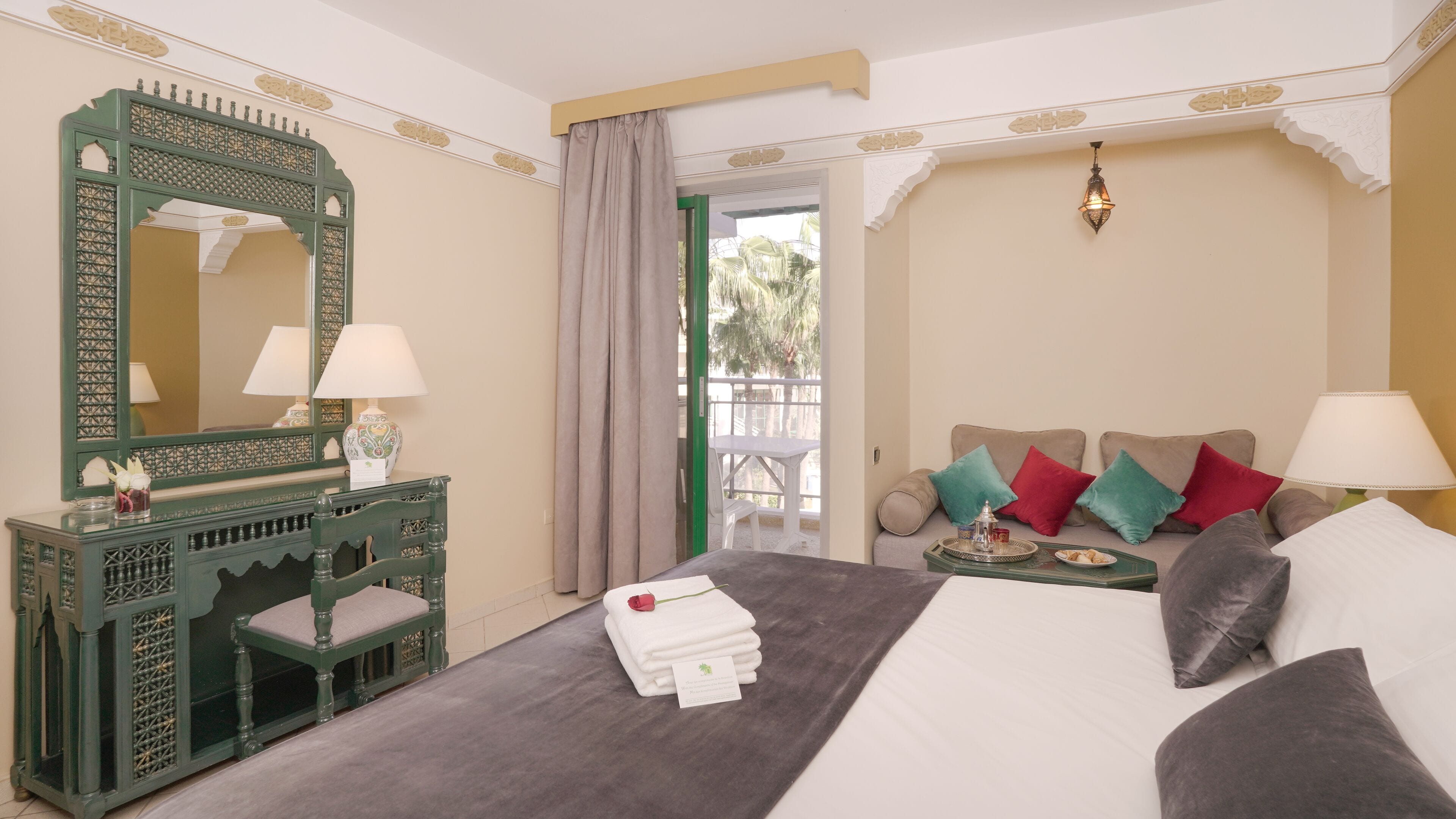 Habitación de hotel con cama, escritorio con espejo, mesita con bandeja y vistas al balcón.