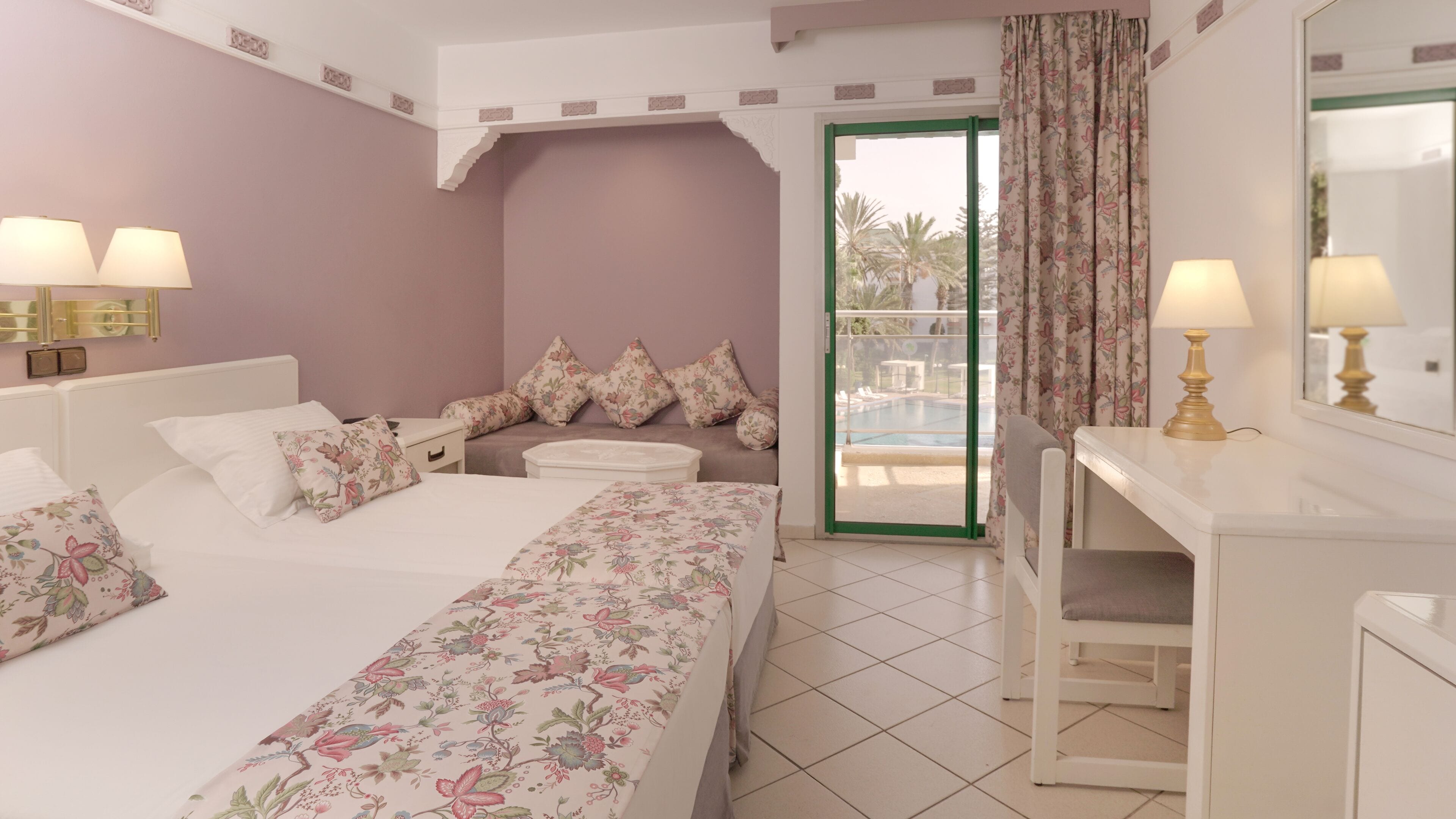 Habitación de hotel con cama, escritorio, silla, mesita de noche, lámpara, espejo y vistas al exterior.