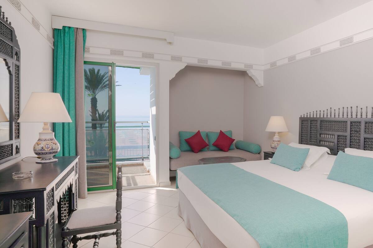 A hotel room with a bed, two bedside tables, a chair, a balcony with a view, and a decorative headboard.