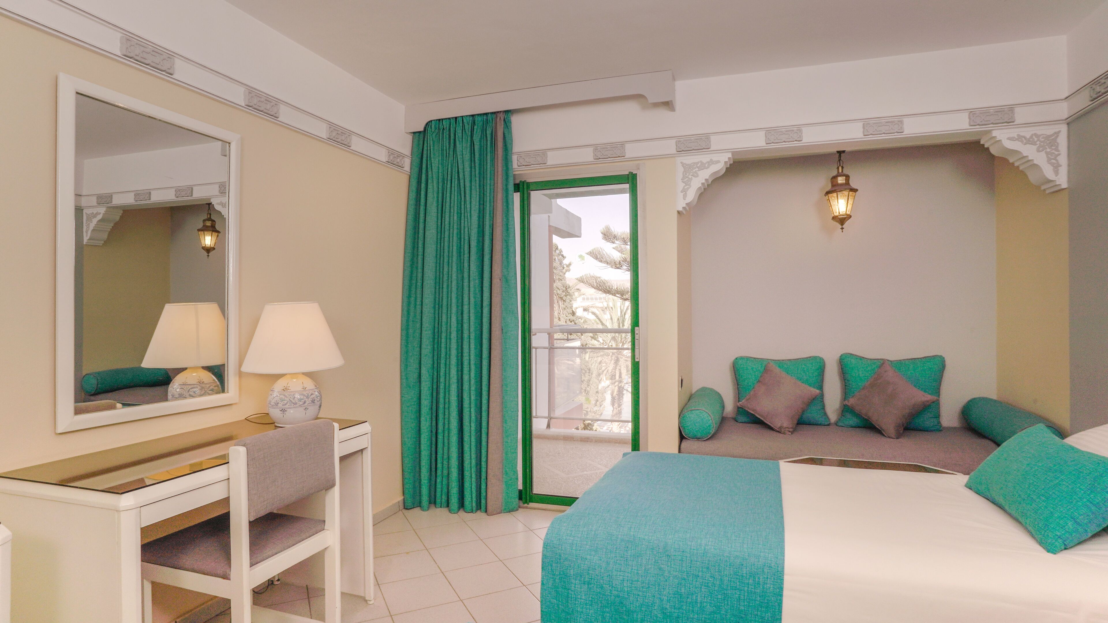 A hotel room with a bed, a desk with a chair, a mirror, a lamp, and a view through a door.