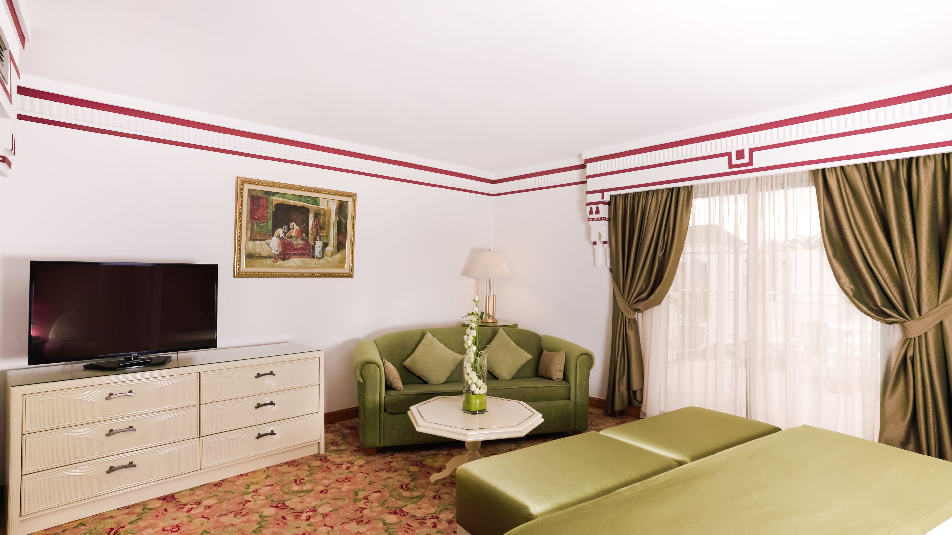 Une chambre d’hôtel avec un canapé vert, un téléviseur à écran plat, une commode, une petite table avec un vase et un tableau accroché au mur.