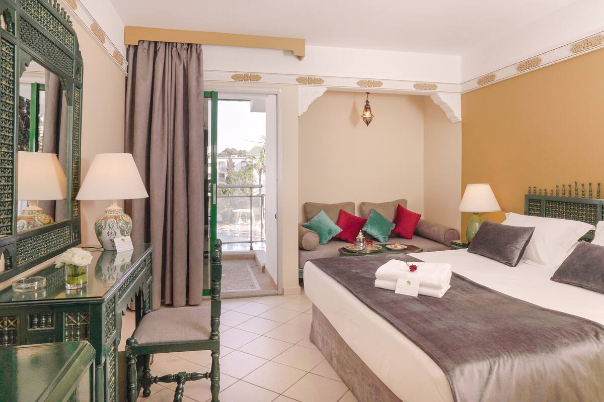 A hotel room with a bed, a desk with a lamp, a chair, a mirror, and a view of the outside.