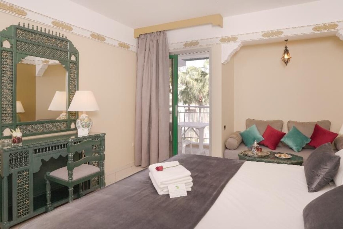 Habitació d'hotel amb un llit, zona de seients amb coixins, tocador amb mirall i vistes al balcó.