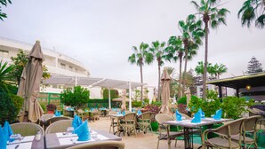 An outdoor dining area with tables and chairs, palm trees, and a building labeled "AGADIR BEACH CLUB HOTEL" in the background. - AGADIR BEACH CLUB HOTEL (Agadir)