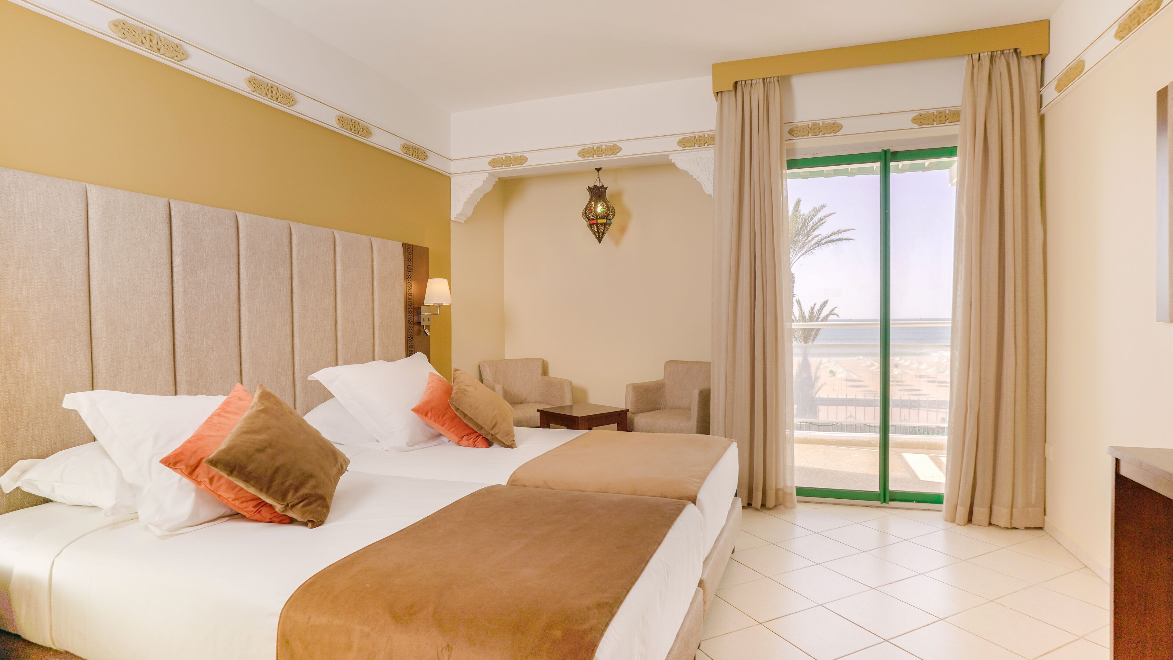 Une chambre d’hôtel avec un lit, deux fauteuils, une petite table, une lampe et une vue sur la plage.