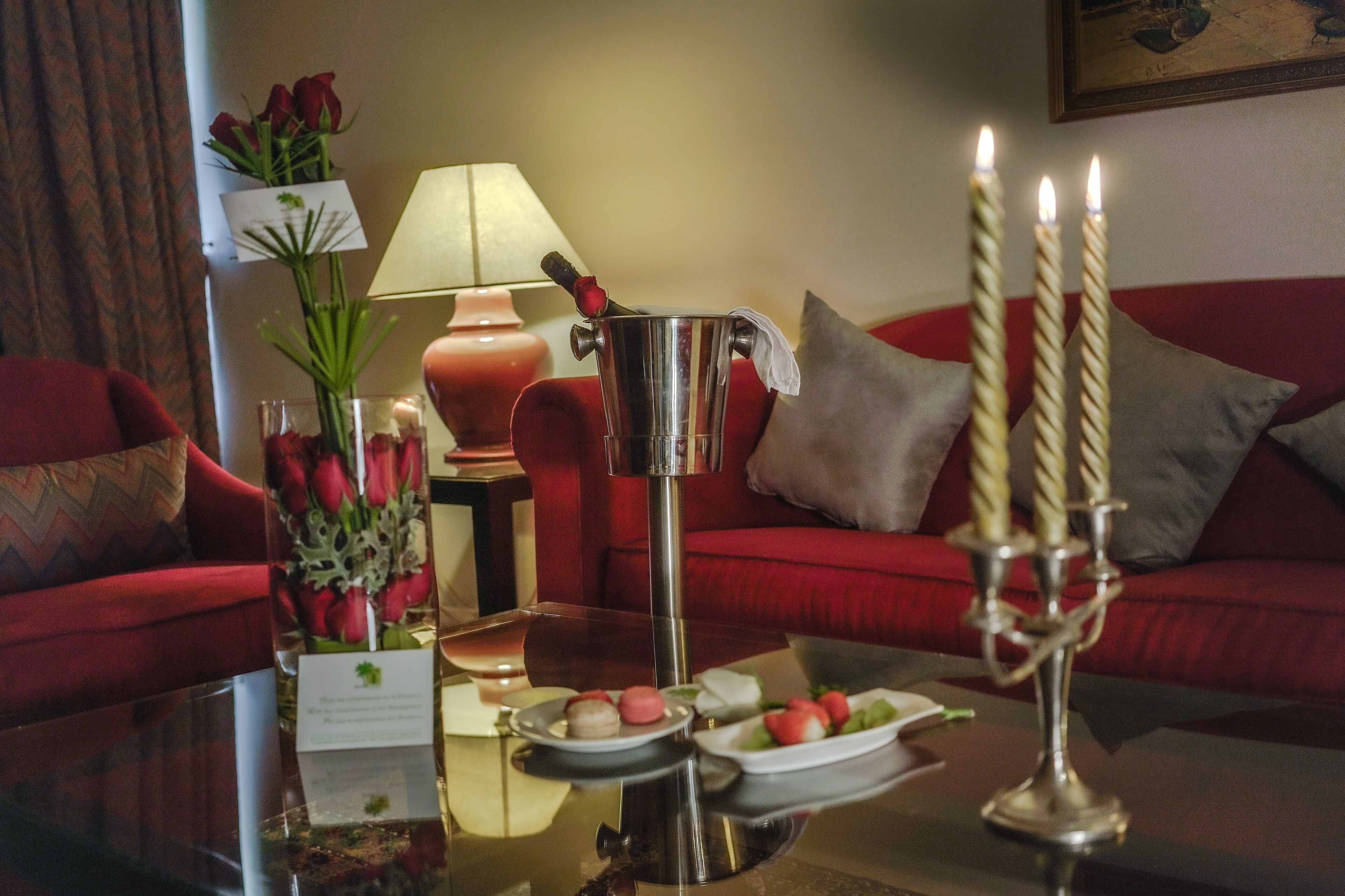 Una sala de estar acogedora con un sofá rojo, una mesa de centro con una jarra de metal, velas y un florero con flores.