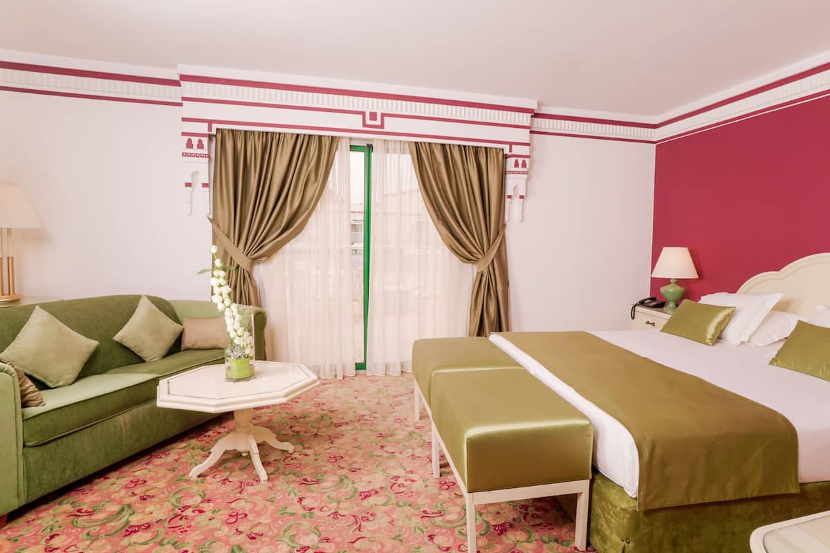 A hotel room with a green sofa, a bed with a green bedspread, a small round table with a lamp, and a window with curtains.