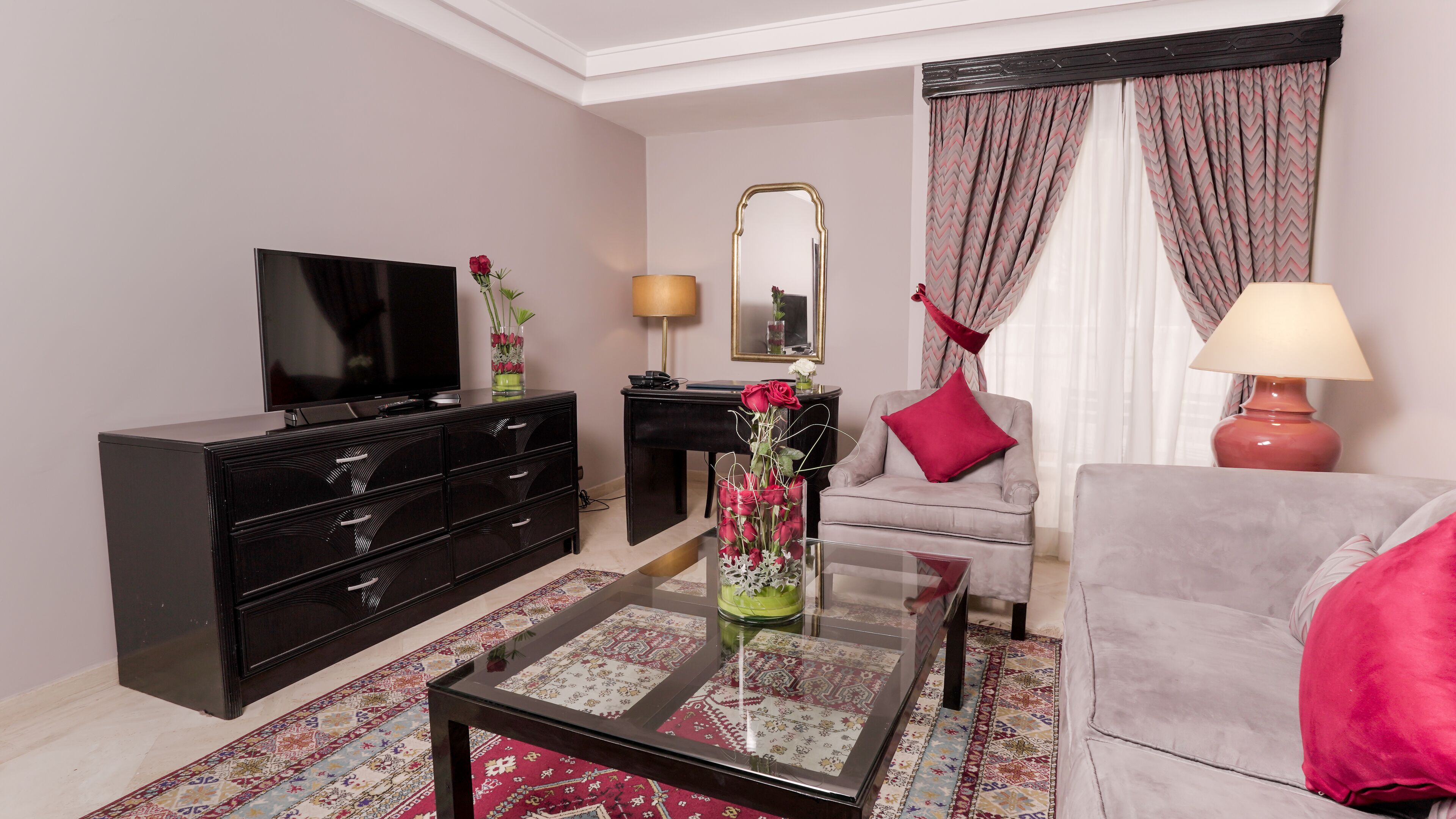 A modern living room with a flat-screen TV, a black dresser, a glass coffee table, a grey sofa, a red chair, a mirror, and a patterned rug.
