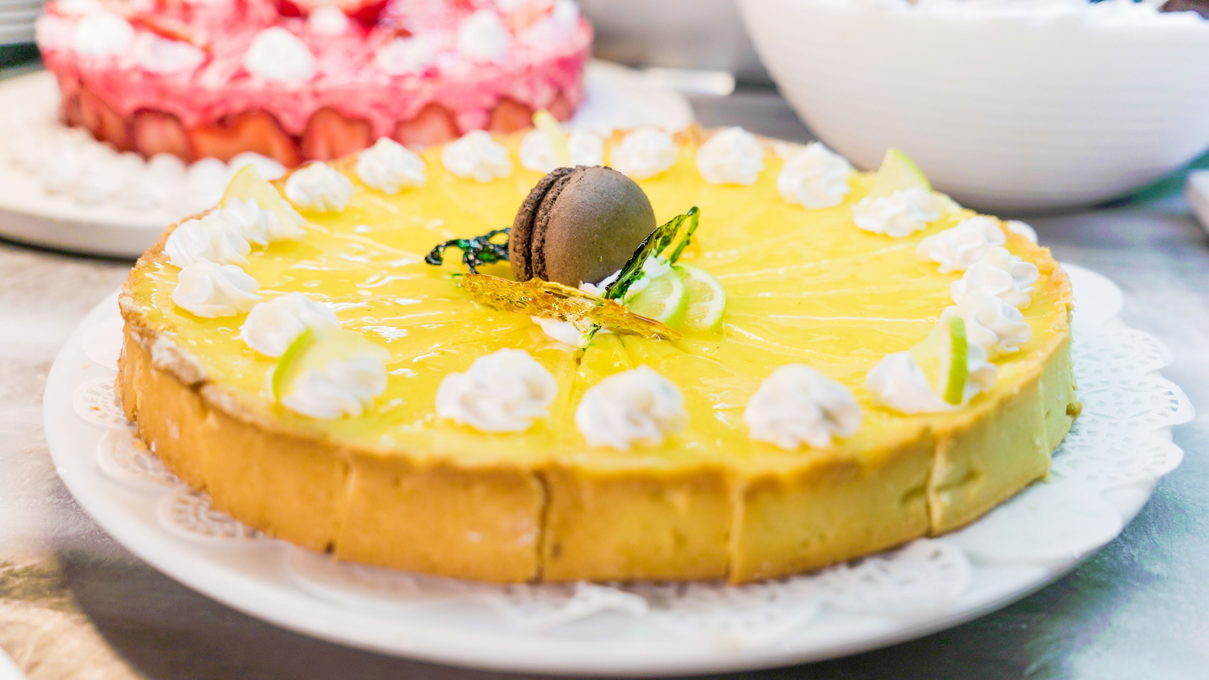 A round yellow cake with white cream dollops and a chocolate macaron on top, placed on a white plate with a lace doily.
