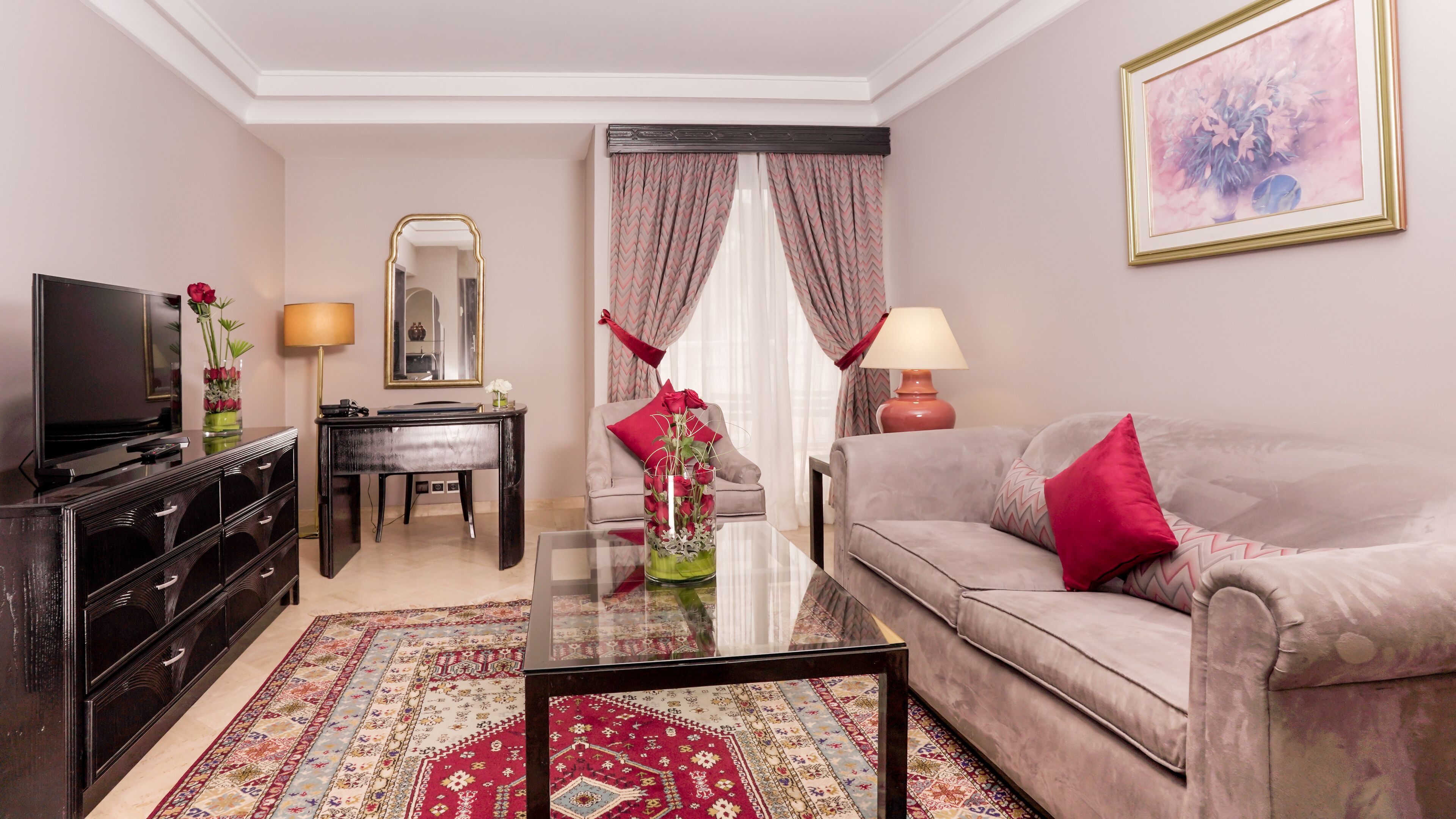 A living room with a sofa, a coffee table, a TV, a desk, and a chair. There is a mirror on the wall and a painting above the sofa.