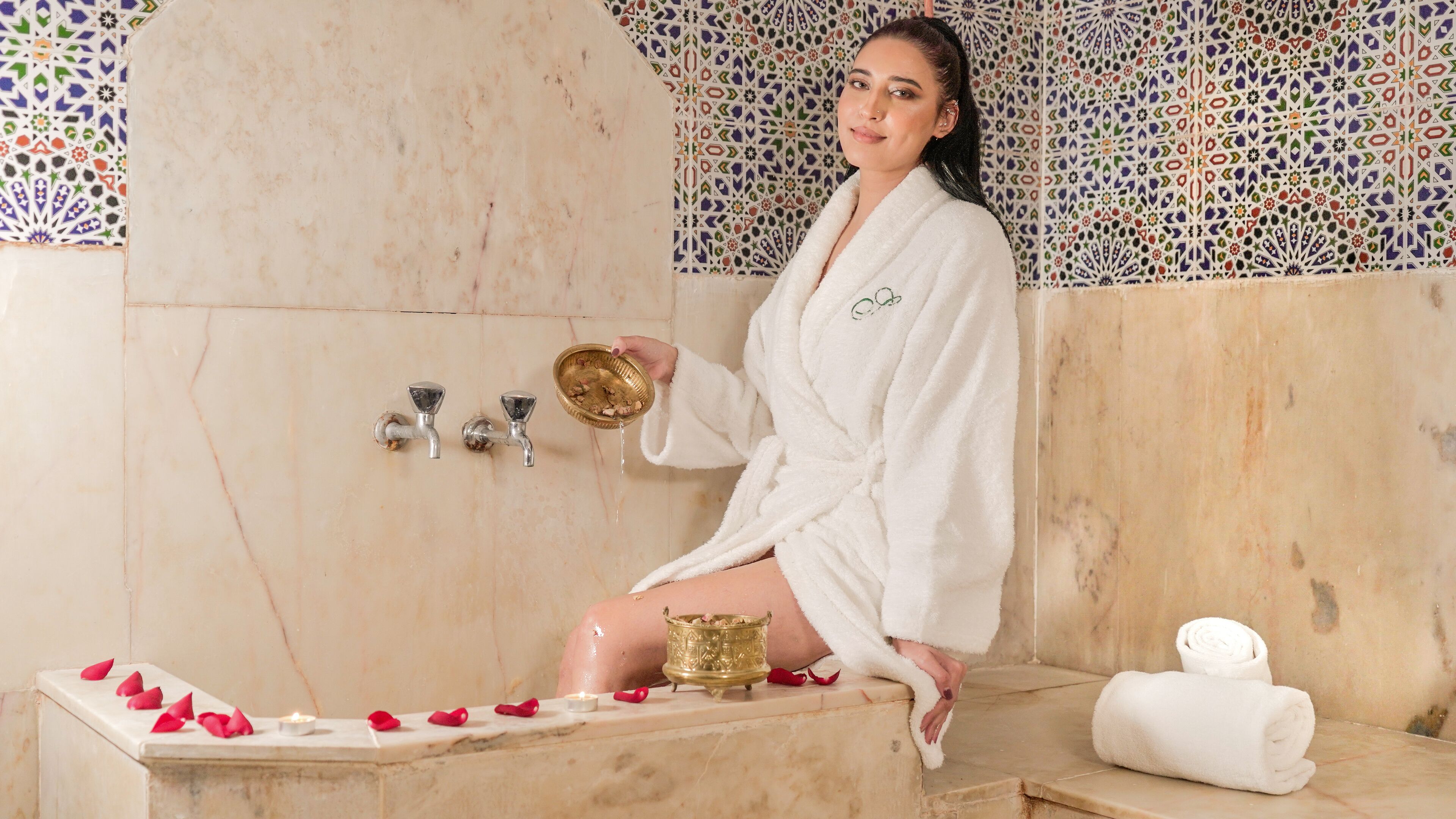 A person in a white bathrobe sitting on a bathtub with rose petals, holding a golden bowl.