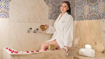 A person in a white bathrobe sitting on a bathtub with rose petals, holding a golden bowl.