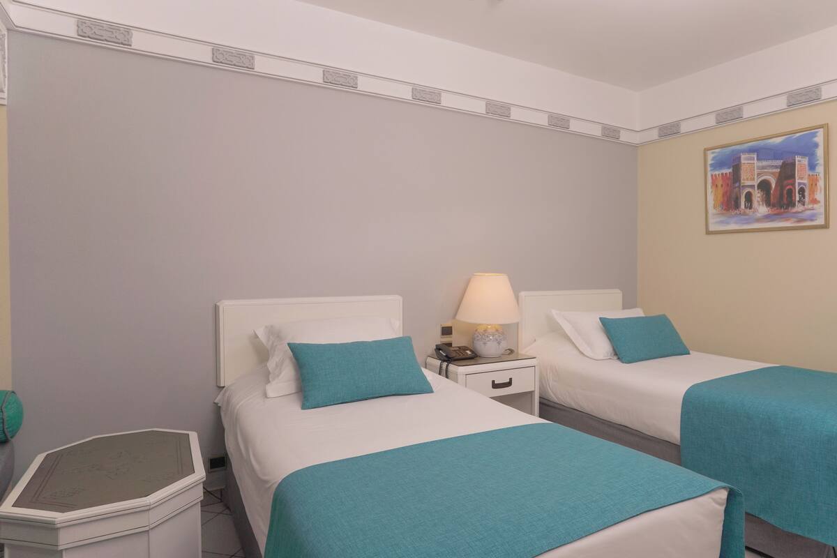 A hotel room with two beds, each with a teal bedspread, a nightstand with a lamp, and a small trash bin.